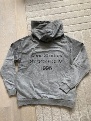 Grå hoodie från Acne Studios S - Snygg grå hoodie från Acne Studios med tryck på ryggen där det står 'Acne Studios STOCKHOLM 1996'. Klassisk passform med huva och ribbade muddar. Perfekt för en chill och stilren look. Materialet känns mjukt och bekvämt.