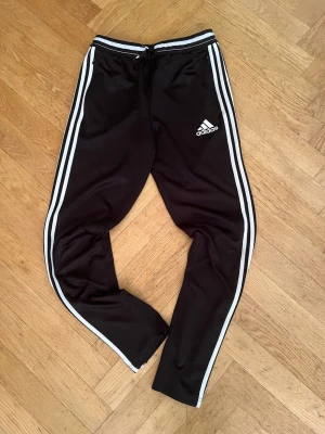 Svarta Adidas climacoo byxor. - adidasModell: Tiro (utrustade med funktionellt ClimaCool-material som andas och transporterar bort fukt)Storlek: 160/164 (motsvarar junior 13–14 år)Färg: Klassiskt svart med de ikoniska tre vita ränderna längs sidorna samt vit broderad logotypDetaljer: Praktiska sidofickor med dragkedja, elastisk midja med dragsko samt smidiga dragkedjor vid anklarna för att enkelt kunna tas av och på över skor.