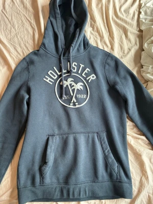 Mörkblå hoodie från Hollister - Mörkblå hoodie från Hollister med stor vit logga och palmtryck på bröstet. Klassisk känguruficka framtill, justerbar huva med dragsko och ribbade muddar. Mjuk bomullsblandning som är skön att ha på sig.