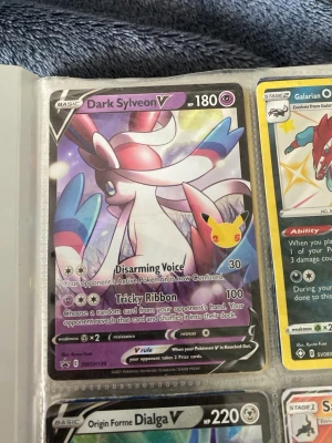 Pokémonkort Dark Sylveon V - Pokémonkortet Dark Sylveon V har en färgstark design i rosa, vitt och blått med holografisk effekt. Kortet har 180 HP och attacker som Disarming Voice och Tricky Ribbon. Perfekt för dig som samlar eller spelar Pokémon TCG.