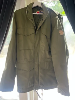 Grön vindjacka från Fjällräven - Snygg grön vindjacka från Fjällräven med klassisk logga på ärmen. Jackan har flera praktiska fickor framtill, hög krage och dragsko i nederkant. Tillverkad i slitstarkt material som passar perfekt för uteliv och äventyr.