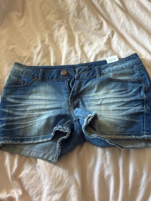 Blå jeansshorts från ONLY - Säljer ett par blå jeansshorts från ONLY med slitna detaljer och fransig kant. Klassisk femficksmodell med knapp och dragkedja framtill. Perfekta för varma dagar och har en avslappnad, casual vibe. De är lågmidjade men den har en slit i bak alltså som ett hål som man ser på sista bilden 