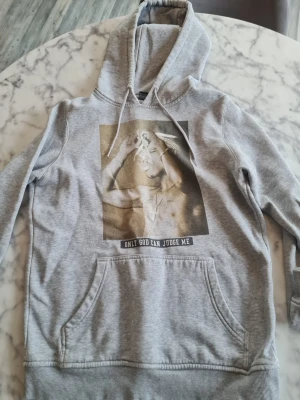 Grå hoodie Mister Tee Tupac S - Säljer en grå hoodie från Mister Tee i storlek S med en stor Tupac-print på bröstet och texten 'Only God Can Judge Me'. Hoodien har huva med dragsko och en stor magficka. Perfekt för dig som gillar streetwear och vill sticka ut.