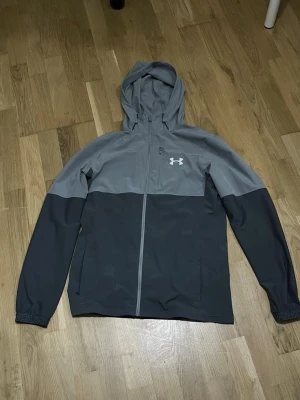 Grå vindjacka från Under Armour - Snygg tvåtonad grå vindjacka från Under Armour med huva och hel dragkedja framtill. Jackan har två sidofickor med dragkedja och logga på bröstet. Perfekt för blåsiga dagar och sportiga outfits.
