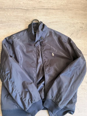 Ralph Lauren jacka - Snygg mörkblå bomberjacka från Polo Ralph Lauren med klassisk broderad logga på bröstet. Jackan har ribbade muddar vid ärmslut och nederkant, ståkrage med knapp och dragkedja framtill. Tillverkad i glansigt material som ger en modern look.