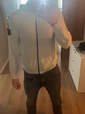 Vit stickad hoodie med dragkedja - Vit stickad hoodie från Stone Island med dragkedja framtill och ribbade muddar. Klassisk patch med logga på vänster ärm. 