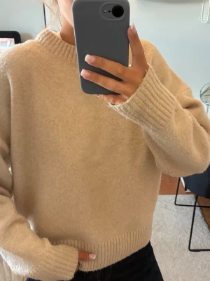 Beige stickad tröja med rund hals - Mysig beige stickad tröja med rund halsringning och ribbade muddar vid ärmslut och nederkant. Tröjan har en avslappnad passform och är perfekt för lager på lager. Materialet ser mjukt och fluffigt ut, vilket ger en skön känsla.