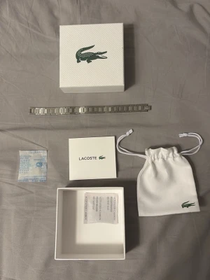 Lacoste silverfärgad klockarmband - Snyggt silverfärgat klockarmband från Lacoste i rostfritt stål. Armbandet har en stilren design med rutmönstrade länkar och klassiskt spänne med Lacoste-loggan. Perfekt för dig som vill byta ut eller uppgradera din klocka med en fräsch och modern look.