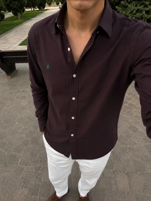 Mörk vinröd skjorta från Ralph Lauren - Säljer en mörk vinröd skjorta från Ralph Lauren med klassisk krage och knappar framtill. Skjortan har lång ärm och är tillverkad i mjuk bomull. Liten grön logga broderad på bröstet för en clean look. 