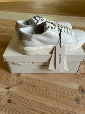Vita sneakers från Calvin Klein - Stilrena vita sneakers från Calvin Klein med tjock vit sula och grå detalj på hälen. Skorna har klassisk snörning och är tillverkade i slätt läder. Perfekta för dig som gillar en clean och modern look.
