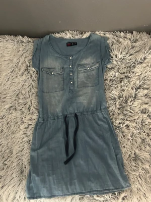 Blå jeansklänning med knytband - Snygg blå jeansklänning med korta ärmar, två bröstfickor och knappar framtill. Midjan har ett svart knytband som ger en avslappnad look. Perfekt för dig som gillar en enkel men trendig stil.