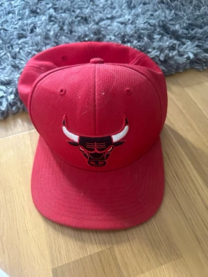 Röd Chicago Bulls keps Mitchell & Ness - Snygg röd snapback-keps från Mitchell & Ness med Chicago Bulls-logga broderad framtill. Kepsen har platt skärm och justerbar baksida för perfekt passform. Tillverkad i bomullstyg och har klassisk streetwear-vibe.