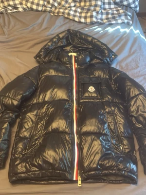 Svart dunjacka från Moncler med huva - Snygg svart dunjacka från Moncler med glansig finish, huva och dragkedja med rödvit detalj. Jackan har en bröstficka med lock och Moncler-logga samt två sidofickor. Perfekt för kalla vinterdagar och riktigt stilren look. Modell Montbeliard