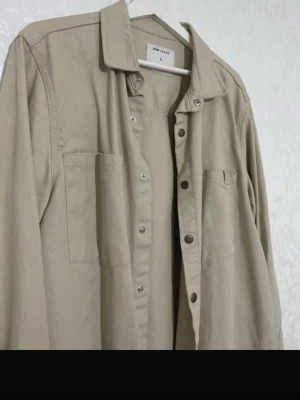 Beige overshirt från Low Jeans - Stilren beige overshirt från Low Jeans i storlek L. Skjortan har två bröstfickor, tryckknappar framtill och på ärmsluten samt klassisk krage. Tillverkad i ett kraftigare bomullstyg som ger en avslappnad och trendig look.