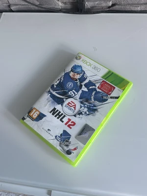 NHL 12 Xbox 360 spel - NHL 12 till Xbox 360, ett ishockeyspel från EA Sports. Omslaget är vitt med blå detaljer och visar en hockeyspelare i action. Spelet kommer i ett klassiskt grönt Xbox 360-fodral och innehåller både skiva och manual.