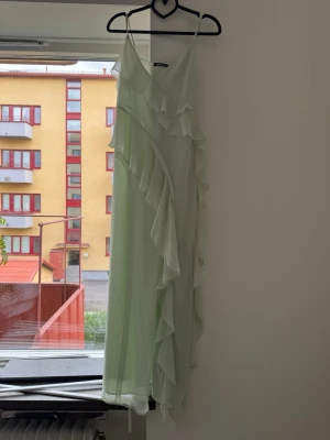 Ljusgrön långklänning med volanger - Säljer en ljusgrön långklänning med tunna axelband och vackra volangdetaljer längs hela klänningen. Klänningen har omlottkänsla och är tillverkad i ett lätt, flödande material som passar perfekt till sommaren.