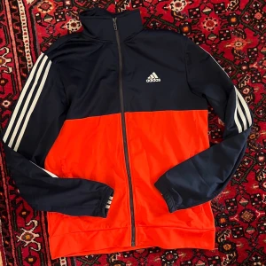 Adidas träningsöverdel marinblå/orangeröd - Snygg träningsöverdel från Adidas i marinblått och orangerött med klassiska vita ränder på ärmarna och Adidas-logga på bröstet. Jackan har dragkedja framtill, hög krage och är tillverkad i ett lätt, sportigt material. Perfekt för träning eller chill.