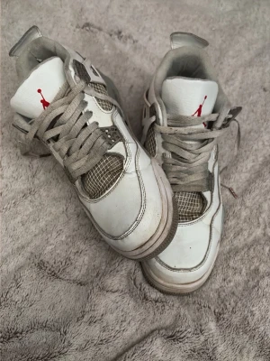 Vita Jordan 4 Oreo sneakers - Säljer ett par klassiska vita Jordan 4 Oreo sneakers med grå mesh-detaljer och röda loggor på plösen. Skorna har högt skaft, tjock sula och ikoniska plastdetaljer på sidorna. Perfekta för dig som gillar streetwear och sportig stil. Skorna har lite sprickor här o där, samt använda mycket. Säljs billigt. Storlek 45 men lite små i storleken. OBS ingen låda till skorna.