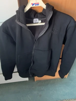 Svart hoodie från C.P. Company - Säljer en svart hoodie från C.P. Company med dragkedja framtill och stor ficka på magen. Hoodien har broderad logga på bröstet och en rejäl huva. Tillverkad i mjukt och bekvämt material, perfekt för en avslappnad stil.