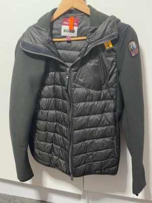 Mörkgrön pufferjacka Parajumpers S - Snygg mörkgrön pufferjacka från Parajumpers i storlek S. Jackan har quiltad front och rygg i glansigt material, stickade ärmar och axlar samt dragkedja framtill. Märkespatch på ärmen och cool detalj vid kragen. Perfekt för kalla vinterdagar.