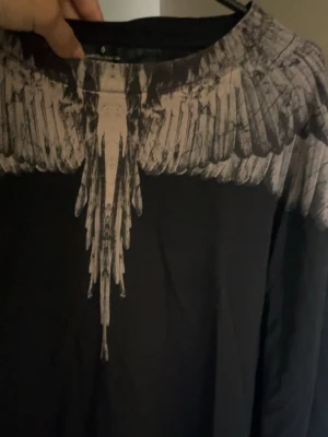Svart tröja med vingmönster  - Säljer en svart tröja från Marcelo Burlon med ikoniskt vingmönster i beige över axlar och bröst. Tröja har rund hals och är tillverkad i mjuk bomull. Perfekt för dig som vill sticka ut med en unik design.