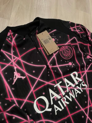 PSG x Jordan svart träningsjacka - Säljer en svart PSG träningsjacka från Jordan med rosa och vita grafiska linjer och prickar. 