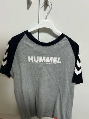 Grå och svart Hummel t-shirt storlek S - Snygg grå t-shirt från Hummel med svarta ärmar och vita detaljer på ärmarna. Stor Hummel-logga framtill. Klassisk rund hals och korta ärmar. Perfekt för en sportig look.