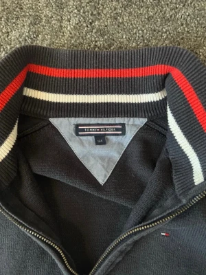 Mörkblå Ziptröja Tommy Hilfiger - Mörkblå ziptröja från Tommy Hilfiger med ribbad krage och muddar. Tröjan har en liten broderad logga på bröstet och snygga ränder i rött och vitt på insidan av kragen. Perfekt till en avslappnad och stilren look. Storlek 158/164 14-15 år