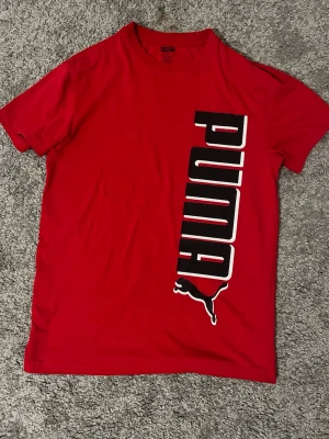 Röd Puma t-shirt med stor logga - Säljer en röd t-shirt från Puma med stor vertikal logga i vitt och svart på framsidan. Klassisk rund hals och korta ärmar. Tillverkad i mjuk bomull som känns skön mot huden. Perfekt för dig som gillar sportig och avslappnad stil.