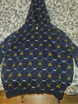 Mörkblå McDonald's x Snipes hoodie - Unik mörkblå hoodie från Snipes i samarbete med McDonald's. Hoodien har ett all-over tryck med gula McDonald's-loggor och vita linjer. Klassisk känguruficka och justerbar huva med snörning. Perfekt för dig som gillar streetwear och vill sticka ut.