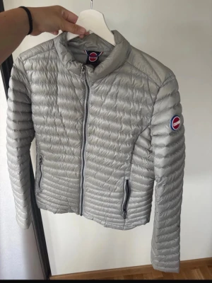 Ljusgrå pufferjacka från Colmar - Snygg ljusgrå pufferjacka från Colmar med quiltad design och hög krage. Jackan har två dragkedjeförsedda fickor och en logotyp på ärmen. Tillverkad i lätt, glansigt material som ger en modern look. Perfekt för kalla dagar. Storleken är M/L. 