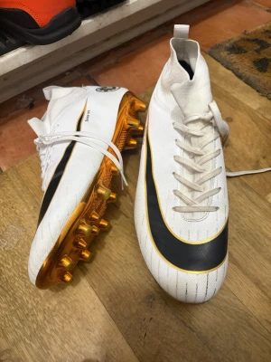 Vita fotbollsskor med guldsula - Säljer ett par vita fotbollsskor med svart och gulddetaljer. Skorna har snörning, högre skaft och en guldfärgad sula med dobbar. Perfekta för dig som vill sticka ut på planen med stilrena och sportiga skor.kan diskutera pris 