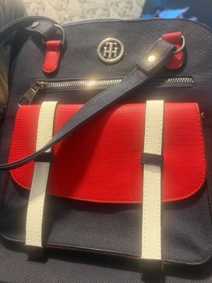 Marinblå och röd ryggsäck Tommy Hilfiger - Snygg marinblå ryggsäck från Tommy Hilfiger med röd frontficka, vita detaljer och metallogo framtill. Väskan har justerbara axelband, dragkedja upptill och en praktisk ytterficka med spännen. Perfekt för dig som vill ha en stilren och sportig look.