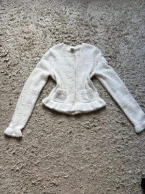 Vit fluffig stickad kofta - Supersöt vit stickad kofta med fluffig känsla och volangkant nedtill. Koftan har långa ärmar, rund halsringning och små fickor framtill. Stängs med knappar och har en mjuk, mysig struktur som känns lyxig och trendig. Den här en tråd som är lös på innersidan av ena ärmen men som inte syns med tröja på.💕