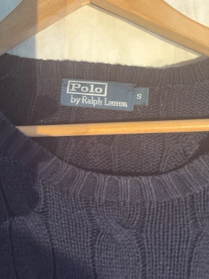 Mörkblå kabelstickad tröja Polo Ralph Lauren - Klassisk mörkblå kabelstickad tröja från Polo Ralph Lauren med rund halsringning och röd broderad logga på bröstet. Tröjan har ribbade muddar vid ärmslut och nederkant, perfekt för en stilren och tidlös look.