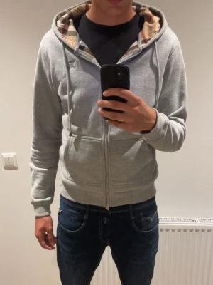 Burberry Grå zip hoodie  - Snygg grå hoodie med dragkedja och huva. Insidan av huvan har ett klassiskt beige, rött och svart rutigt mönster. Tröjan har fickor framtill och ribbade muddar vid ärmar och midja. Perfekt för en avslappnad och trendig look. Storlek L men passar även M!