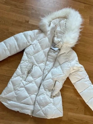 Vit dunjacka med fuskpälsluva - Snygg vit dunjacka med quiltat mönster och dragkedja framtill. Jackan har två fickor med dragkedja och en stor huva med fluffig fuskpäls. Perfekt för kalla vinterdagar och ger en stilren look.