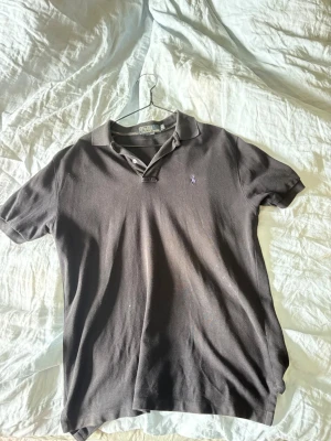 Svart pikétröja från Polo Ralph Lauren - Klassisk svart pikétröja från Polo Ralph Lauren med krage och knappar framtill. Tröjan har en liten broderad logga på bröstet och är tillverkad i bomull för en bekväm känsla. Perfekt till jeans eller shorts.
