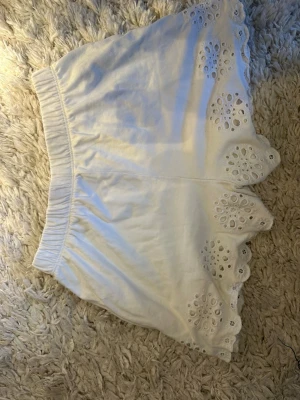Vita shorts med broderade detaljer Zara - Säljer ett par vita shorts från Zara med elastisk midja och fina broderade detaljer längs bensluten. Shortsen har en lös passform och är perfekta för varma dagar. Materialet känns mjukt och luftigt.