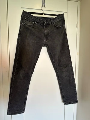 Mörkgrå raka jeans - Säljer ett par mörkgrå jeans med rak passform och klassisk femficksdesign. Jeansen har svarta sömmar och subtila slitningar för en avslappnad look. Perfekta till en streetwear-outfit eller för en enkel vardagsstil.