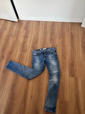 Blå slitna jeans Replay Anbass - Säljer ett par blå jeans med slitningar och raka ben. Jeansen har klassisk femficksdesign, ljus tvätt och detaljerade slitningar framtill och baktill. Materialet är denim och de har bälteshällor samt knappgylf. Pris diskuterbart vid snabb affär.