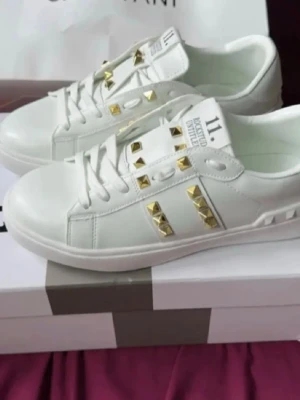 Vita sneakers med gulddetaljer - Säljer ett par vita sneakers med guldfärgade nitar och snörning. Skorna har en stilren design med dekorativa detaljer på sidan och ovandelen. Tillverkade i skinnliknande material och passar perfekt till en trendig streetstyle-look.