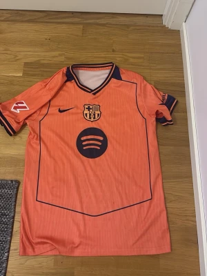 Orange FC Barcelona fotbollströja - Säljer en orange FC Barcelona fotbollströja med mörkblå detaljer och klubbmärke på bröstet. Tröjan har korta ärmar, V-ringning och är tillverkad i ett lätt syntetmaterial som passar perfekt till träning eller match.