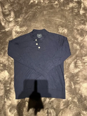 Mörkblå stickad tröja Grand Frank - Snygg mörkblå stickad tröja från Grand Frank med tre knappar vid halsen och ribbade muddar. Tröjan har en klassisk rak passform och är tillverkad i mjukt material som passar perfekt till olika outfits sitter som xs