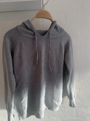 Grå Kashmir hoodie - Säljer en stilren grå stickad hoodie med huva och dragsnören. Hoodien har långa ärmar och ribbade muddar vid ärmslut och nederkant. Perfekt för dig som gillar en enkel och clean look.