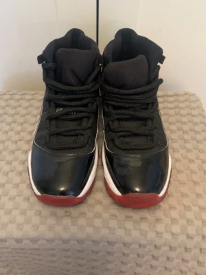 Jordan 11 - Snygga höga sneakers i svart med röda och vita detaljer. Skorna har en blank finish på tån, svart mesh på ovandelen och röda yttersulor. Klassisk siluett med snörning och vadderad plös. Perfekta för dig som gillar streetwear och sportig stil.