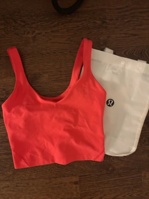 Korall topp från Lululemon - Snygg korall croppad linnetopp från Lululemon med breda axelband och djup rundad halsringning. Perfekt för träning eller som ett statement-plagg till vardags. Mjukt och stretchigt material som sitter bekvämt mot kroppen.