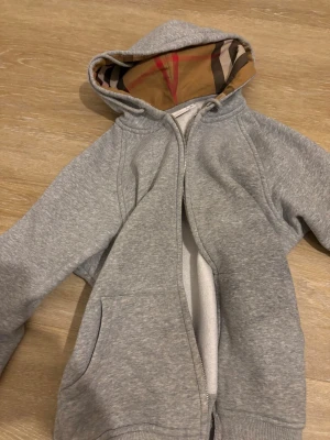 Grå hoodie med dragkedja Burberry - Snygg grå hoodie från Burberry med dragkedja och fickor framtill. Huvan har det klassiska Burberry-mönstret på insidan och tröjan har en diskret broderad logga på bröstet. Perfekt för en avslappnad och trendig look.