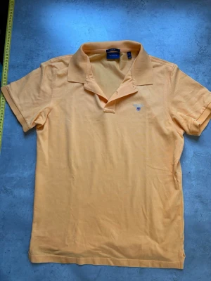 Orange pikétröja från Gant - Klassisk gul pikétröja från Gant med broderad logga på bröstet. Tröjan har korta ärmar, krage och knappslå framtill. Tillverkad i bomull för en skön och luftig känsla. Perfekt till jeans eller shorts för en avslappnad look.