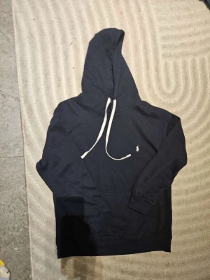 Marinblå hoodie från Polo Ralph Lauren - Snygg marinblå hoodie från Polo Ralph Lauren med vit dragsko och klassisk logga broderad på bröstet. Hoodien har huva och ribbade muddar vid ärmslut och nederkant. Perfekt för en avslappnad och stilren look.